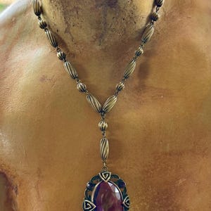 May include: A vintage-style necklace with a bronze-toned chain featuring oval and ribbed beads. The pendant showcases a large, oval, deep purple gemstone, framed by a decorative bronze setting with smaller purple accents.