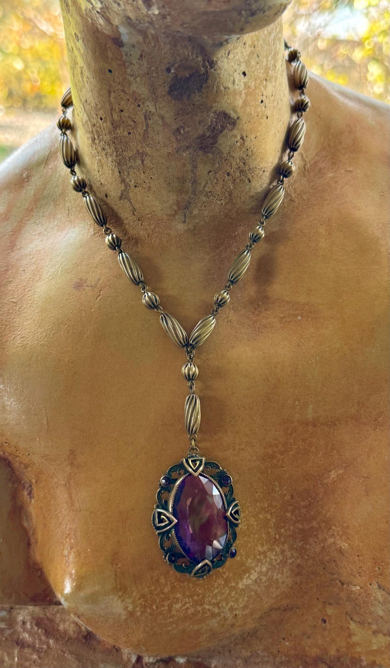 May include: A vintage-style necklace with a bronze-toned chain featuring oval and ribbed beads. The pendant showcases a large, oval, deep purple gemstone, framed by a decorative bronze setting with smaller purple accents.