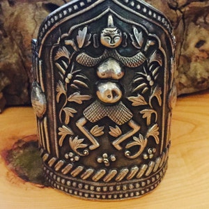 Puede incluir: Un brazalete de plata con un diseño intrincado que presenta una figura humana estilizada rodeada de motivos florales. El brazalete tiene una parte superior redondeada y una superficie texturizada.