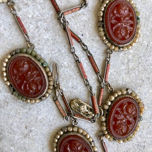 May include: An antique necklace featuring oval, reddish-brown carved cameos with floral designs. The cameos are set in silver-toned metal and surrounded by small, off-white beads. The necklace has a delicate chain with red and silver accents.
