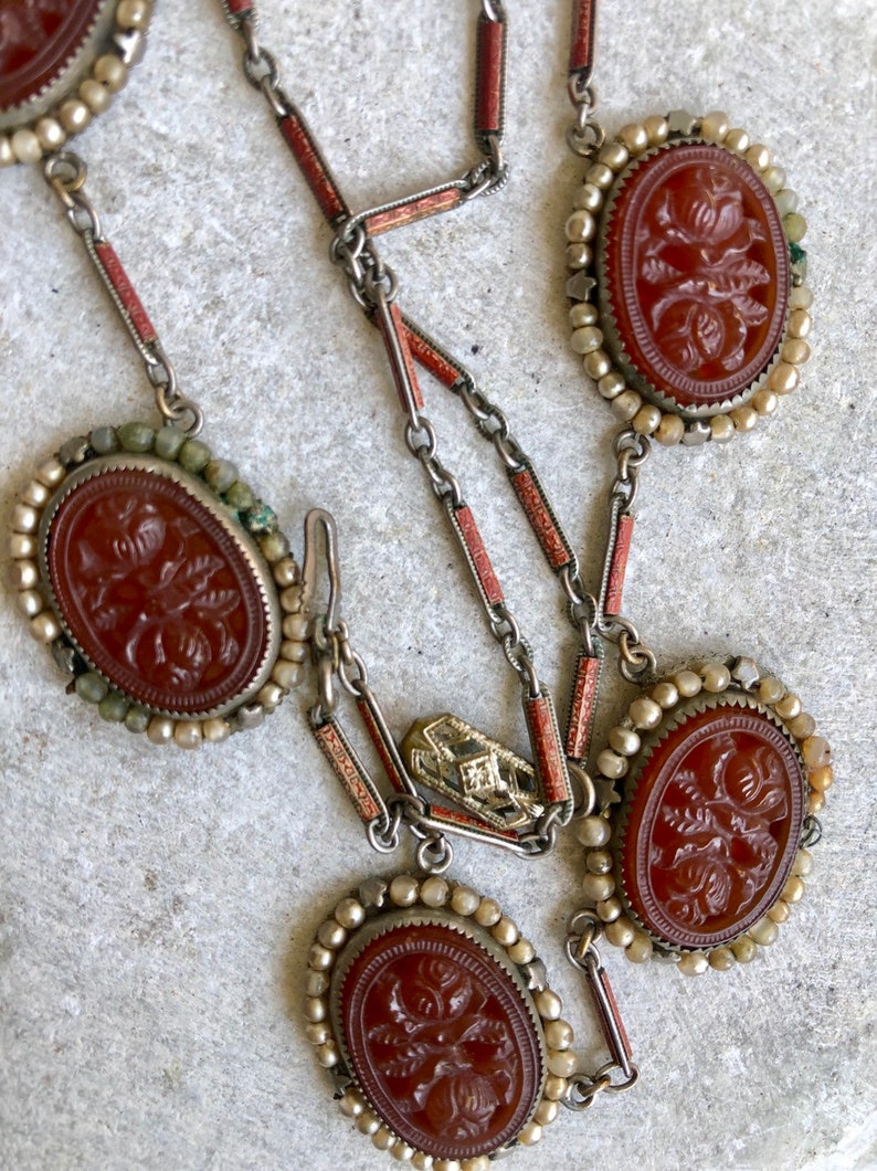 May include: An antique necklace featuring oval, reddish-brown carved cameos with floral designs. The cameos are set in silver-toned metal and surrounded by small, off-white beads. The necklace has a delicate chain with red and silver accents.