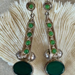 Pu&ograve; includere: Un paio di orecchini color argento con pietre preziose verdi. Ogni orecchino presenta una grande pietra verde ovale nella parte inferiore, con pietre verdi pi&ugrave; piccole ed elementi decorativi argentati. Gli orecchini sono esposti su uno sfondo strutturato bianco sporco.