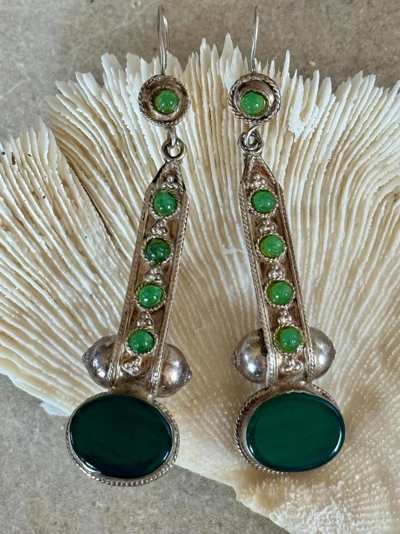 Pu&ograve; includere: Un paio di orecchini color argento con pietre preziose verdi. Ogni orecchino presenta una grande pietra verde ovale nella parte inferiore, con pietre verdi pi&ugrave; piccole ed elementi decorativi argentati. Gli orecchini sono esposti su uno sfondo strutturato bianco sporco.