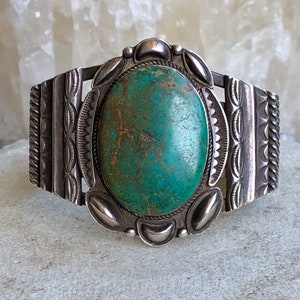 Puede incluir: Una pulsera de plata con una gran piedra ovalada de turquesa engastada en el centro. La pulsera tiene dise&ntilde;os intrincados en los lados.