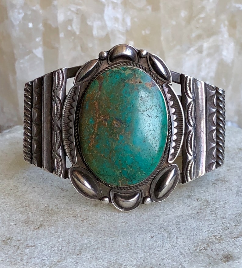 Puede incluir: Una pulsera de plata con una gran piedra ovalada de turquesa engastada en el centro. La pulsera tiene dise&ntilde;os intrincados en los lados.