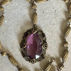 May include: Antique brass-toned necklace featuring an oval purple gemstone pendant. The pendant is framed by an ornate, filigree-style setting with small purple accents. The necklace chain has a mix of ribbed and round beads.