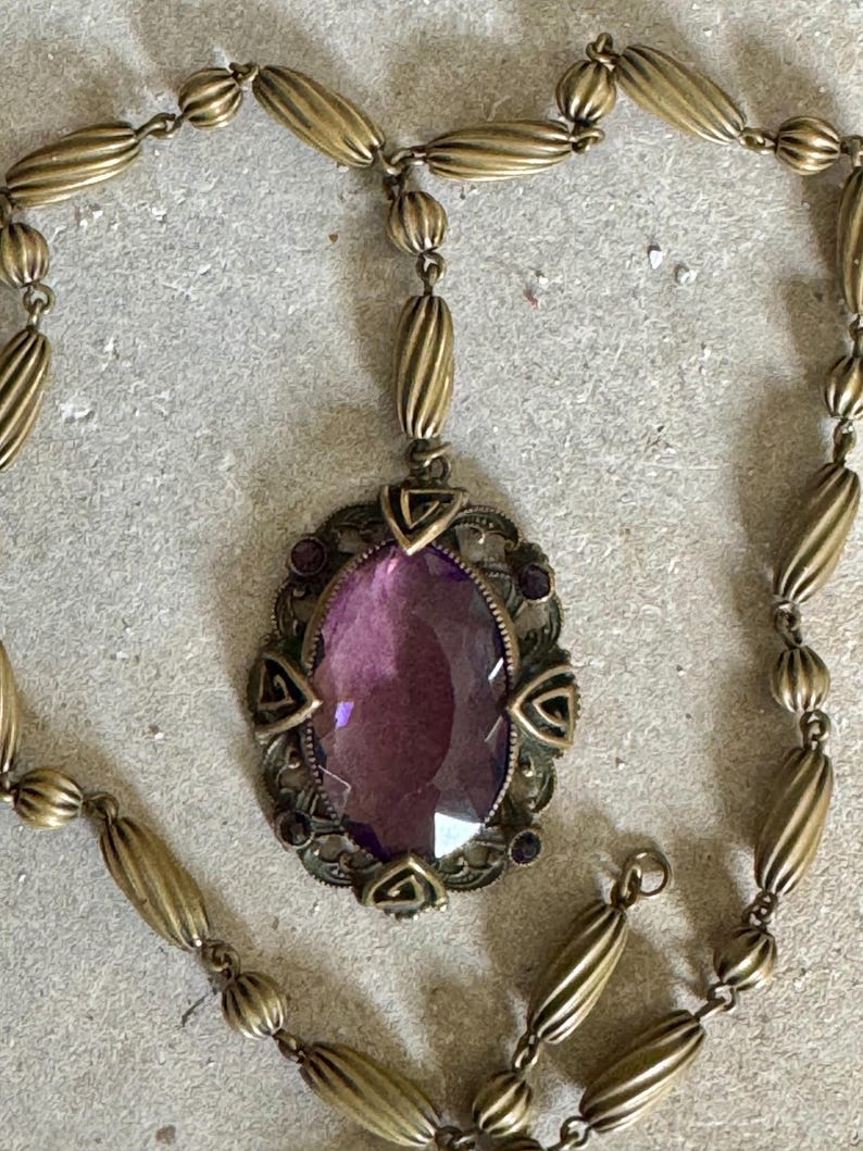 May include: Antique brass-toned necklace featuring an oval purple gemstone pendant. The pendant is framed by an ornate, filigree-style setting with small purple accents. The necklace chain has a mix of ribbed and round beads.