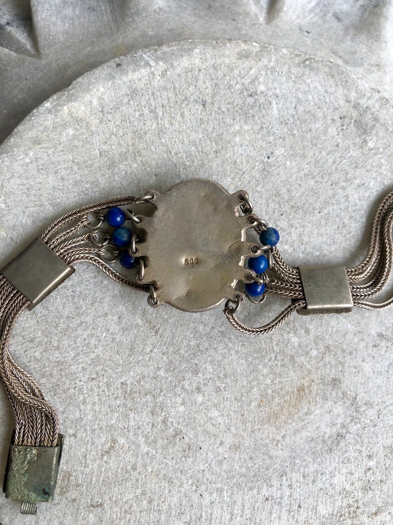 May include: A silver bracelet with a round, ornate charm and blue beads. The charm has the number "900" stamped on it.