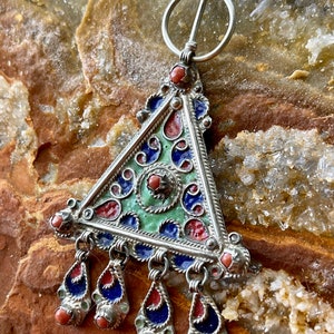 May include: A silver triangular brooch with blue, green, and red enamel and coral accents. The brooch has a decorative design and a pin closure.