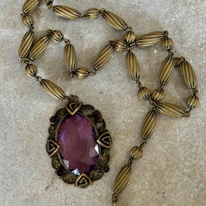 May include: Vintage-style brass necklace featuring an oval, faceted purple pendant. The pendant is framed by a decorative border with small purple accents. The necklace chain is composed of ribbed, oval-shaped beads.