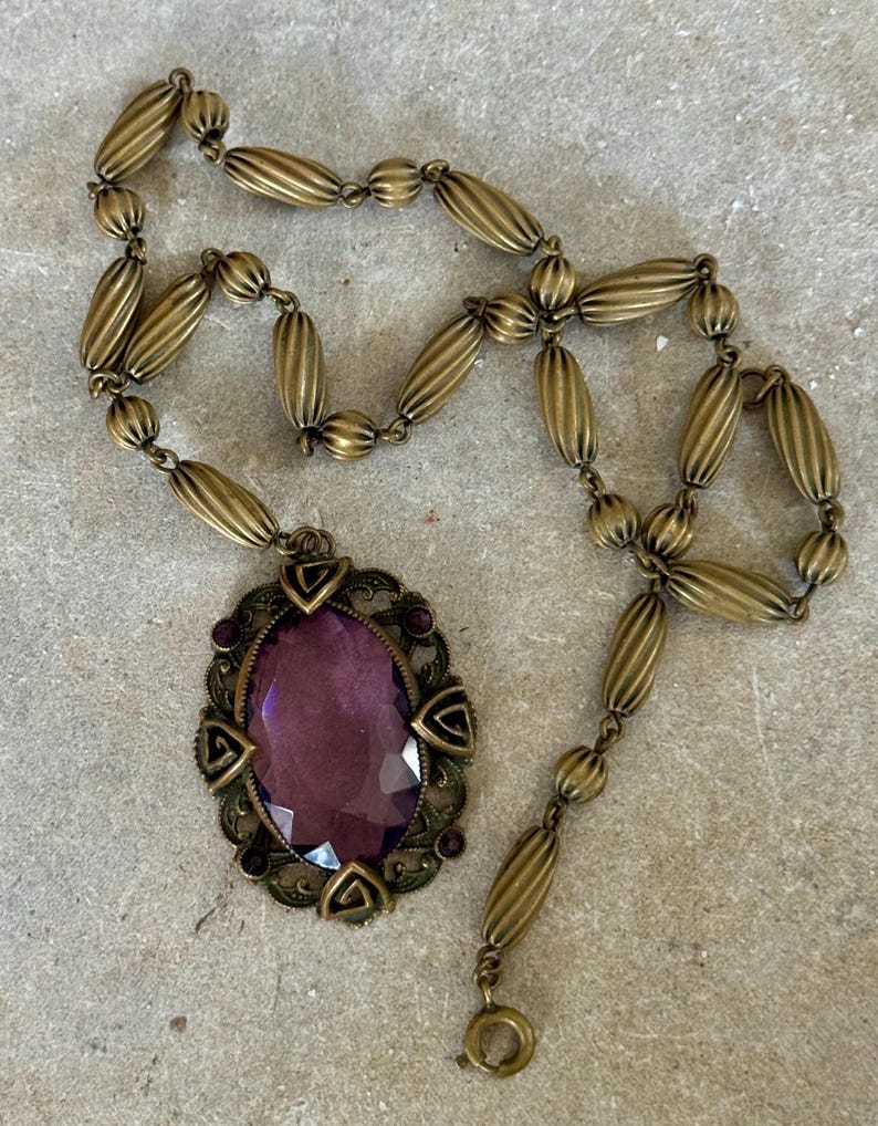 May include: Vintage-style brass necklace featuring an oval, faceted purple pendant. The pendant is framed by a decorative border with small purple accents. The necklace chain is composed of ribbed, oval-shaped beads.