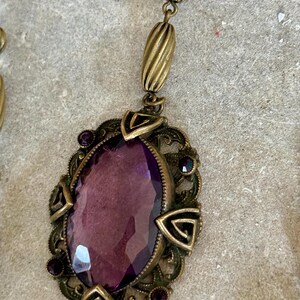 May include: An antique-style necklace with a large, oval, faceted purple gemstone pendant. The pendant is set in a detailed, ornate, bronze-colored metal frame with small purple accent stones. The necklace has a matching bronze chain.