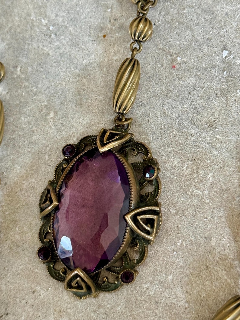 May include: An antique-style necklace with a large, oval, faceted purple gemstone pendant. The pendant is set in a detailed, ornate, bronze-colored metal frame with small purple accent stones. The necklace has a matching bronze chain.
