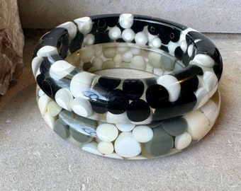 May include: Two black and white bangle bracelets with a pebble design. The bracelets are made of clear resin with embedded black, white, and gray pebbles.