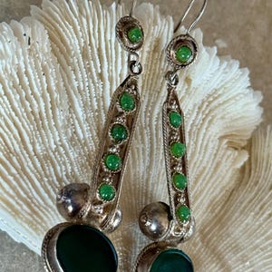 Pu&ograve; includere: Un paio di orecchini pendenti color argento con pietre preziose verdi. Ogni orecchino presenta una grande pietra verde ovale nella parte inferiore, una sfera d'argento e una fila verticale di pietre verdi pi&ugrave; piccole. Chiusura a gancio.