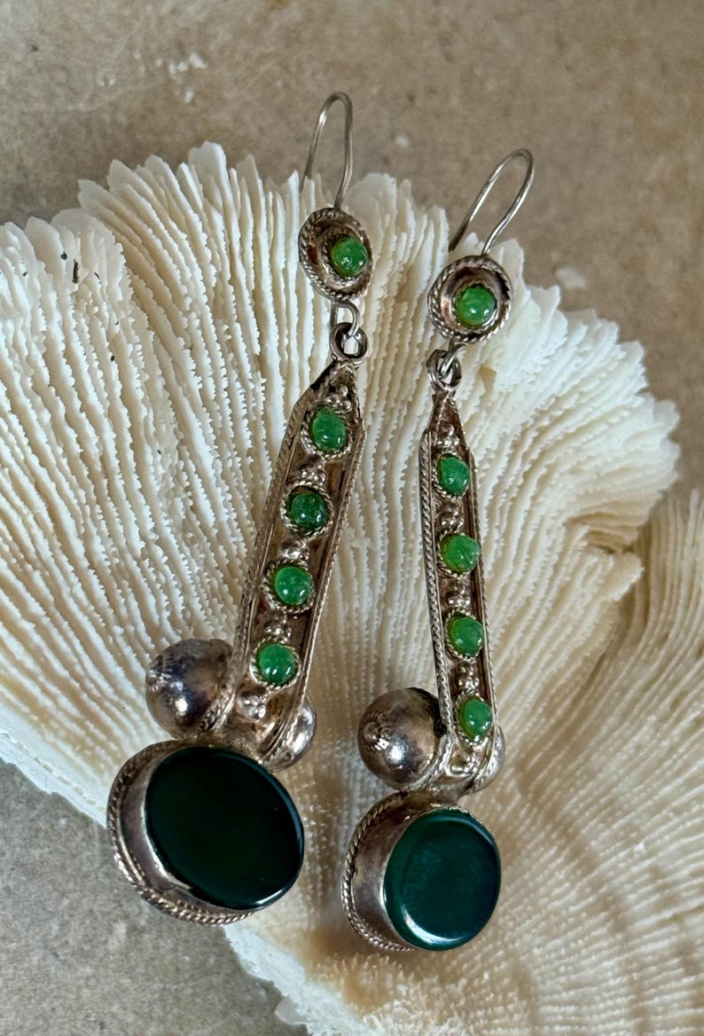 Pu&ograve; includere: Un paio di orecchini pendenti color argento con pietre preziose verdi. Ogni orecchino presenta una grande pietra verde ovale nella parte inferiore, una sfera d'argento e una fila verticale di pietre verdi pi&ugrave; piccole. Chiusura a gancio.