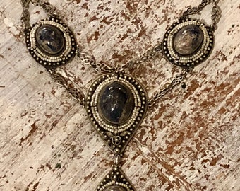 Puede incluir: Un collar de cadena de plata con tres colgantes de forma ovalada. Cada colgante presenta una piedra gris oscura rodeada por un anillo de pequeñas cuentas blancas. El collar está sobre una superficie de madera.