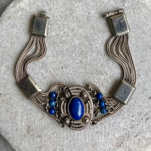 May include: A silver bracelet with a blue lapis lazuli stone set in a silver filigree design. The bracelet has multiple strands of chain and is accented with small blue beads.