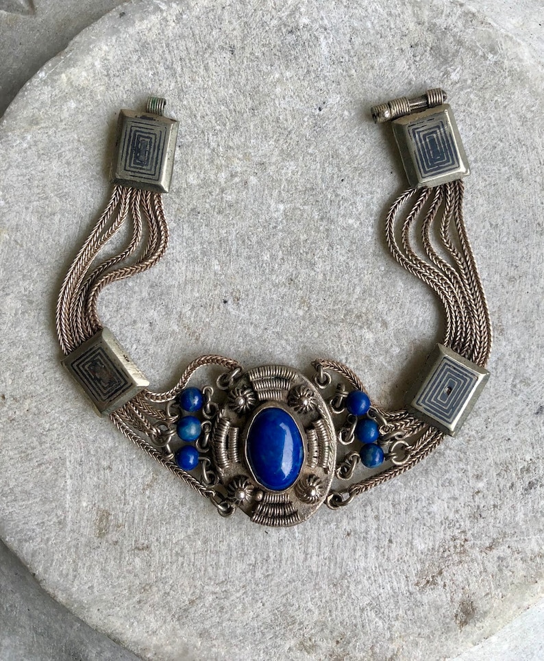 May include: A silver bracelet with a blue lapis lazuli stone set in a silver filigree design. The bracelet has multiple strands of chain and is accented with small blue beads.