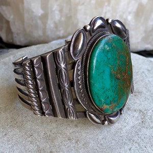 Puede incluir: Un brazalete de plata con una gran piedra ovalada de turquesa engastada en un engaste de plata. El brazalete tiene un dise&ntilde;o decorativo con un patr&oacute;n repetitivo de peque&ntilde;os c&iacute;rculos y l&iacute;neas.