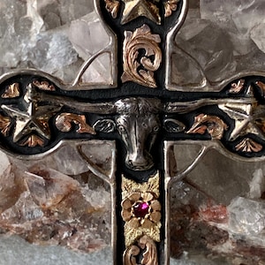 May include: A silver and gold cross pendant featuring a black bull's head at its centre. The cross is adorned with stars and floral designs. A red gemstone is set in the centre of the cross.
