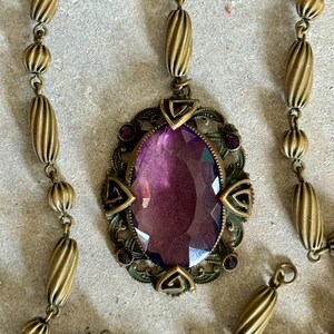 May include: Antique brass-toned necklace featuring an oval pendant with a large, faceted purple gemstone. The pendant is framed by an ornate, filigree design with small purple accents. The chain has a series of ribbed and spherical beads.