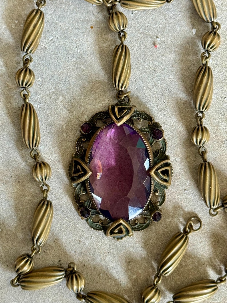 May include: Antique brass-toned necklace featuring an oval pendant with a large, faceted purple gemstone. The pendant is framed by an ornate, filigree design with small purple accents. The chain has a series of ribbed and spherical beads.