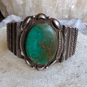 Puede incluir: Un brazalete de plata con una gran piedra ovalada de turquesa engastada en el centro. El brazalete est&aacute; decorado con intrincados dise&ntilde;os de plata.