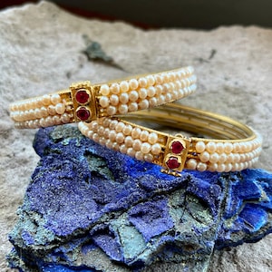 May include: Two gold bangles with rows of white pearls and red gemstone accents.