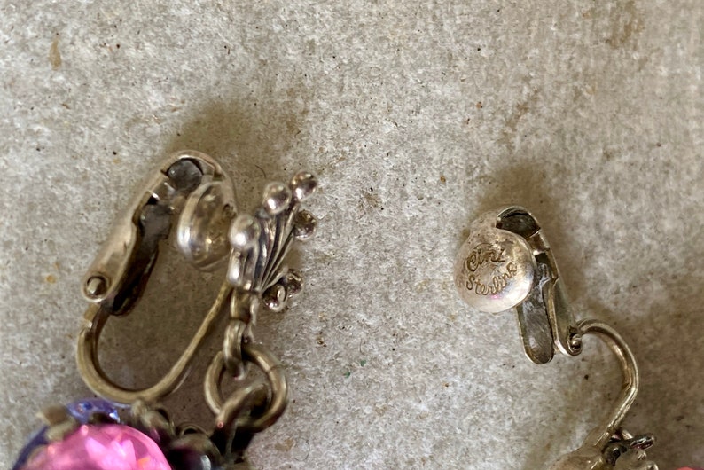 May include: Close-up of a pair of silver clip-on earrings with pink and purple stones. The earrings are marked "925 Sterling".
