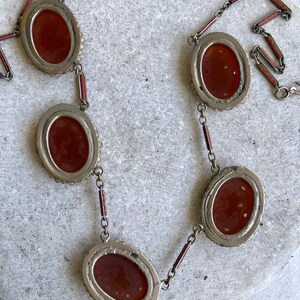 May include: An antique necklace featuring oval, reddish-brown cabochons set in silver-toned metal. The cabochons are linked by small chains and red-painted bars. The necklace is displayed on a light-colored, textured surface.