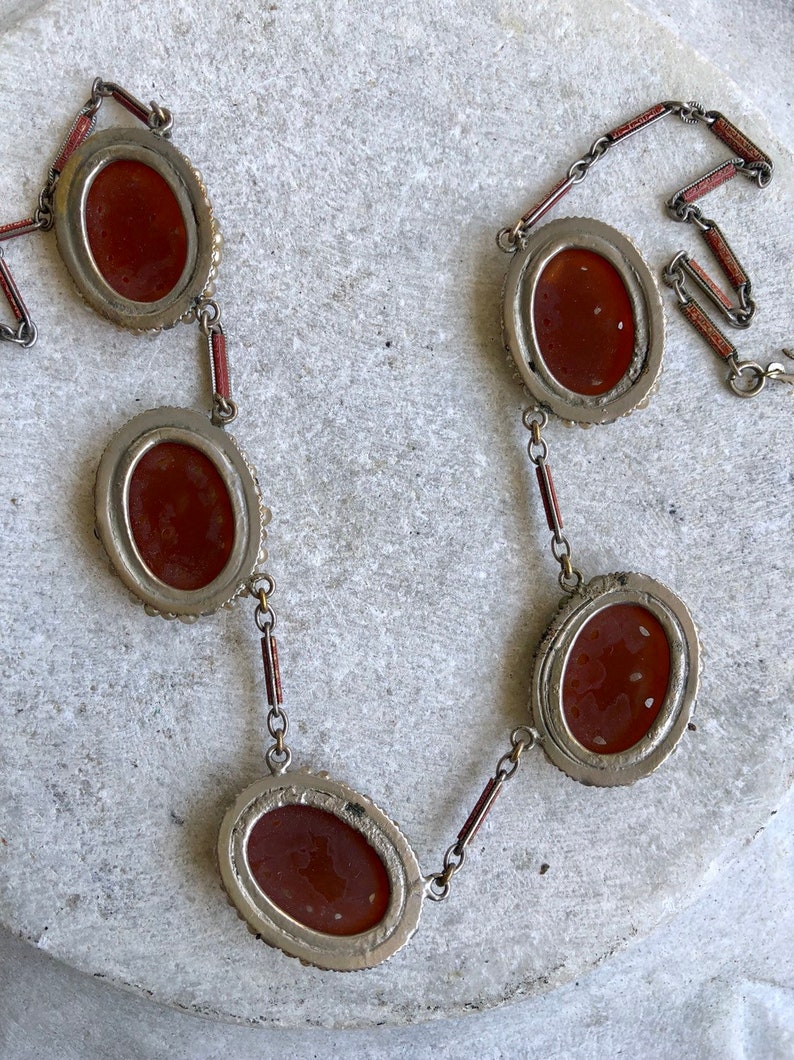 May include: An antique necklace featuring oval, reddish-brown cabochons set in silver-toned metal. The cabochons are linked by small chains and red-painted bars. The necklace is displayed on a light-colored, textured surface.