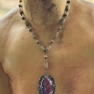 May include: Antique-style necklace with a large, oval, deep purple gemstone pendant. The pendant is set in an ornate, bronze-toned metal frame. The necklace features a beaded chain and a Y-shaped drop design, showcasing vintage elegance.