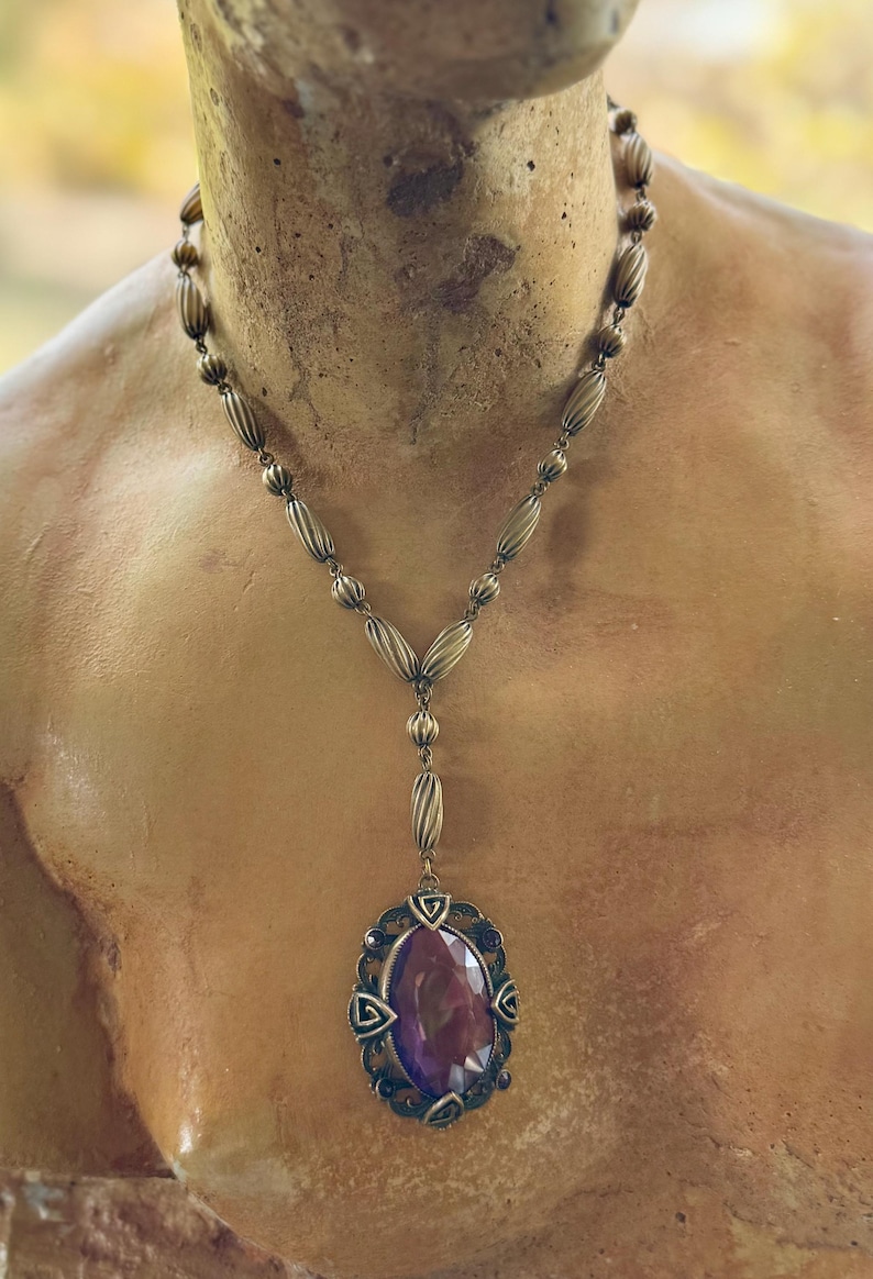 May include: Antique-style necklace with a large, oval, deep purple gemstone pendant. The pendant is set in an ornate, bronze-toned metal frame. The necklace features a beaded chain and a Y-shaped drop design, showcasing vintage elegance.