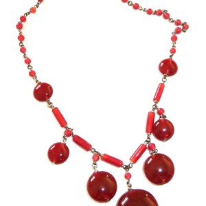 May include: A red glass bead necklace with a silver clasp. The necklace has a variety of red beads, including small round beads and larger, flat, round beads.