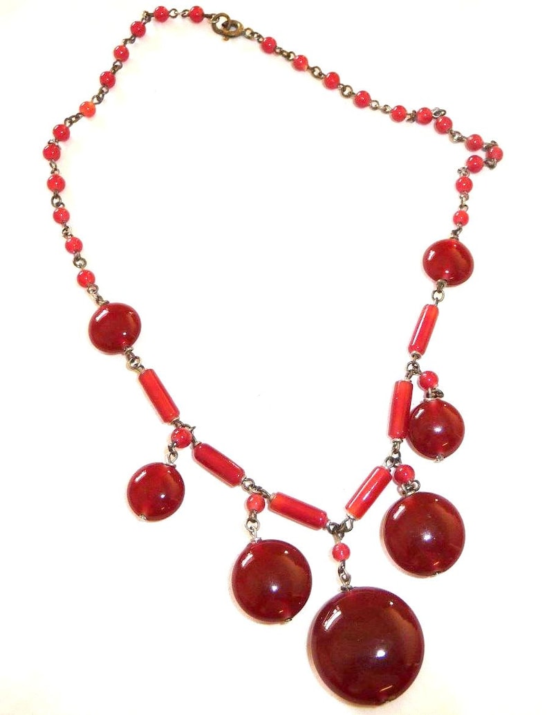 May include: A red glass bead necklace with a silver clasp. The necklace has a variety of red beads, including small round beads and larger, flat, round beads.