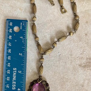 May include: Antique brass-toned necklace with an oval purple gemstone pendant. The pendant is approximately 2 inches long and is surrounded by a decorative filigree frame. The necklace chain features a series of ribbed beads and small round beads.