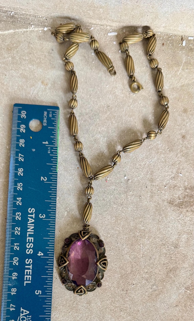 May include: Antique brass-toned necklace with an oval purple gemstone pendant. The pendant is approximately 2 inches long and is surrounded by a decorative filigree frame. The necklace chain features a series of ribbed beads and small round beads.