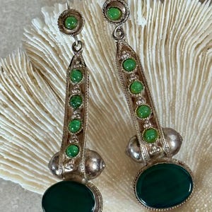 Pu&ograve; includere: Un paio di orecchini pendenti color argento con dettagli verdi. Ogni orecchino presenta una grande pietra verde ovale in basso, una serie di pietre verdi pi&ugrave; piccole e una pietra verde circolare in alto. Gli orecchini hanno un'estetica vintage.