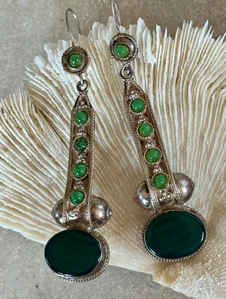 Pu&ograve; includere: Un paio di orecchini pendenti color argento con dettagli verdi. Ogni orecchino presenta una grande pietra verde ovale in basso, una serie di pietre verdi pi&ugrave; piccole e una pietra verde circolare in alto. Gli orecchini hanno un'estetica vintage.