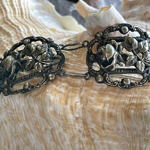May include: A silver bracelet with a delicate floral design. The bracelet is made of linked oval-shaped pieces with intricate cutouts and raised details. The floral design features leaves and flowers.