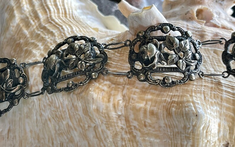 May include: A silver bracelet with a delicate floral design. The bracelet is made of linked oval-shaped pieces with intricate cutouts and raised details. The floral design features leaves and flowers.