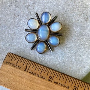 K&ouml;nnte beinhalten: Eine silberne Brosche mit einem Sternburst-Design mit acht schillernden blau-wei&szlig;en Steinen. Die Brosche hat einen Durchmesser von etwa 2,5 cm.