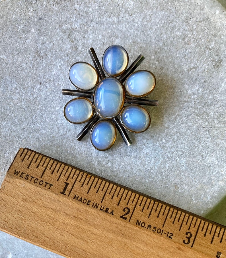K&ouml;nnte beinhalten: Eine silberne Brosche mit einem Sternburst-Design mit acht schillernden blau-wei&szlig;en Steinen. Die Brosche hat einen Durchmesser von etwa 2,5 cm.