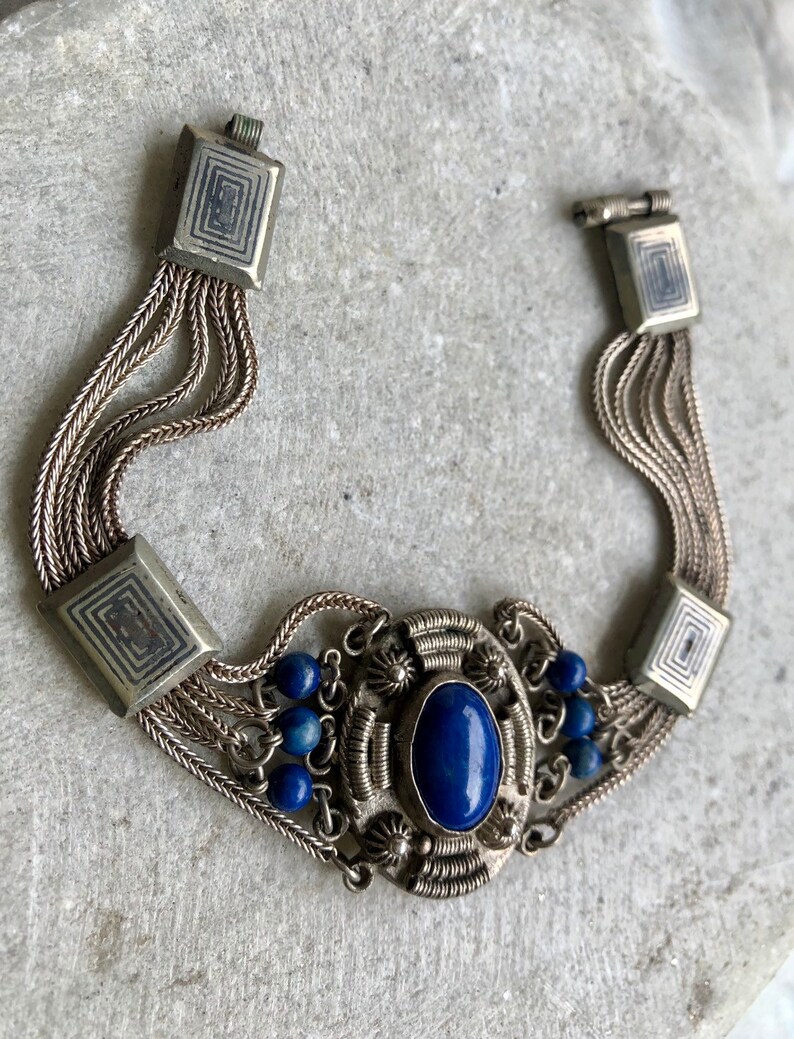 May include: A silver bracelet with a blue lapis lazuli stone and smaller blue beads. The bracelet has a chain-like design and square-shaped clasps.