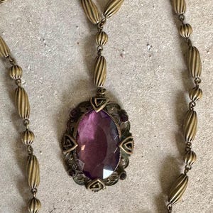 May include: An antique brass-toned necklace featuring an oval, faceted, purple gemstone pendant. The pendant is framed by an ornate, filigree design with small purple accents. The necklace chain has a series of elongated, ribbed beads.