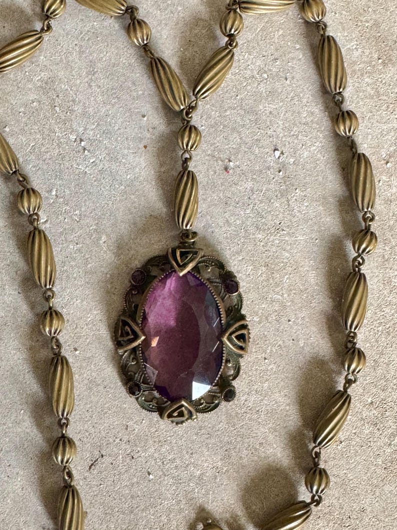 May include: An antique brass-toned necklace featuring an oval, faceted, purple gemstone pendant. The pendant is framed by an ornate, filigree design with small purple accents. The necklace chain has a series of elongated, ribbed beads.
