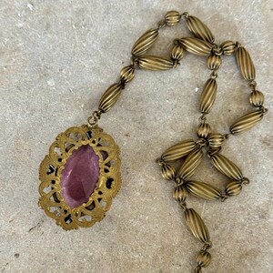May include: Antique brass-toned necklace featuring an oval pendant with a purple center stone. The pendant is framed by an ornate, openwork design. The necklace chain is composed of elongated, ribbed beads.