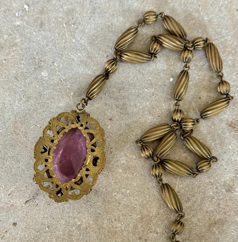 May include: Antique brass-toned necklace featuring an oval pendant with a purple center stone. The pendant is framed by an ornate, openwork design. The necklace chain is composed of elongated, ribbed beads.