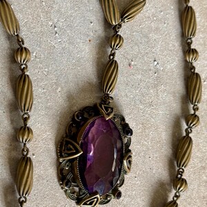 May include: Antique gold-tone necklace featuring an oval, faceted purple gemstone pendant. The pendant is set in an ornate filigree frame with geometric accents. The necklace chain has a series of ribbed and round beads.